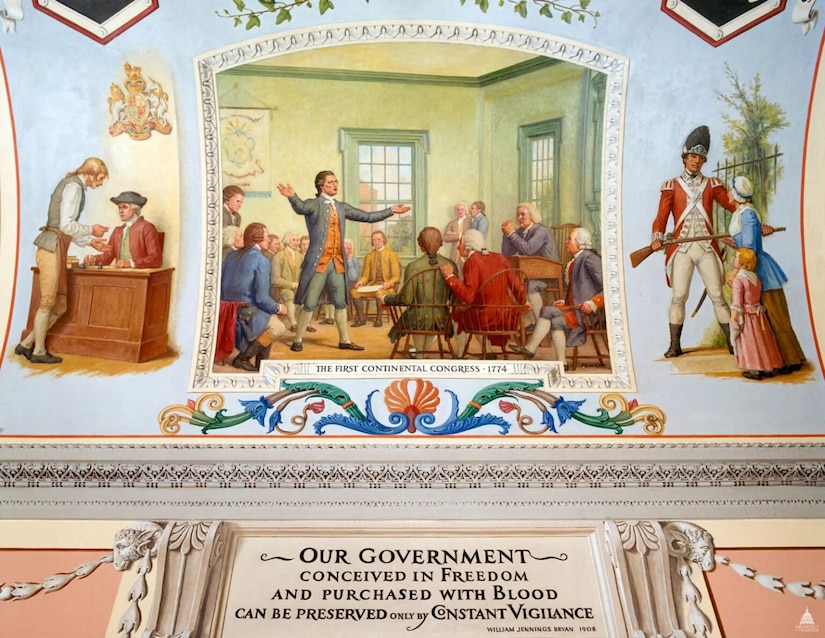 A mural is shown depicting a man in colonial attire standing in front of a small desk where another man in similar attire is seated. In the center of the mural, a man in colonial attire stands with outstretched arms while speaking to other seated men in similar attire. On the right side of the mural, a man in a military uniform with a gun faces a woman and a small girl in colonial attire.