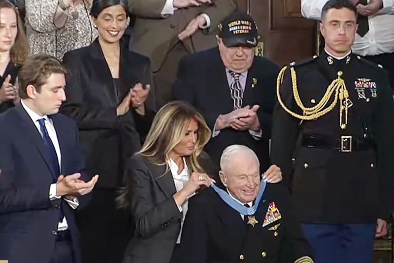 A woman in business attire places a medal around the neck of an elderly man in a military dress uniform, who is seated. Dozens of people in business attire are clapping around them.