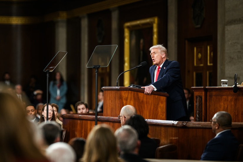 A man in a suit speaks into a microphone while standing behind a lectern. In front of him are dozens of seated people.