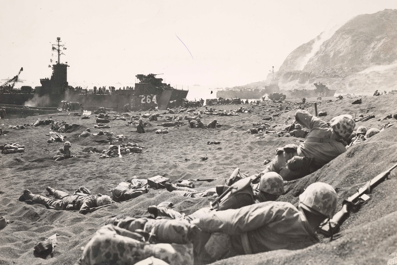 Hundreds of helmet-clad men lie on sand and in foxholes dug into a beach. In the distance, about three ships sit near the shore.