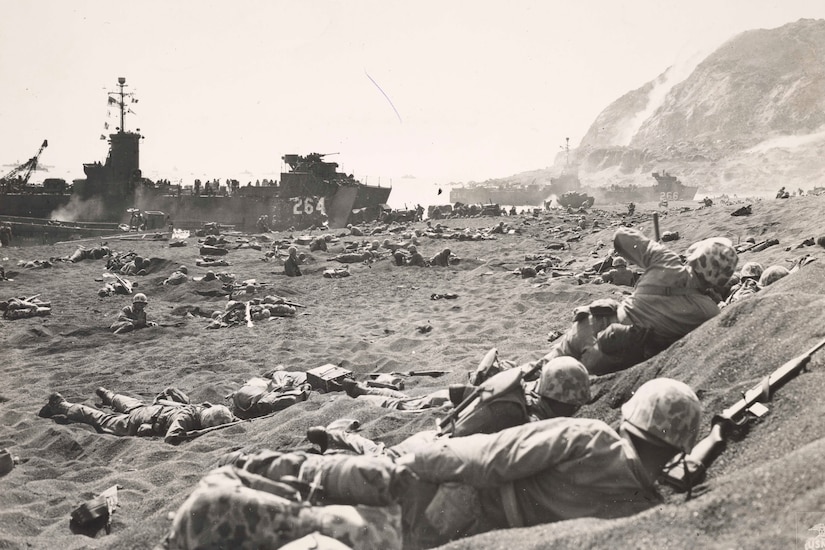 Hundreds of helmet-clad men lie on sand and in foxholes dug into a beach. In the distance, about three ships sit near the shore.