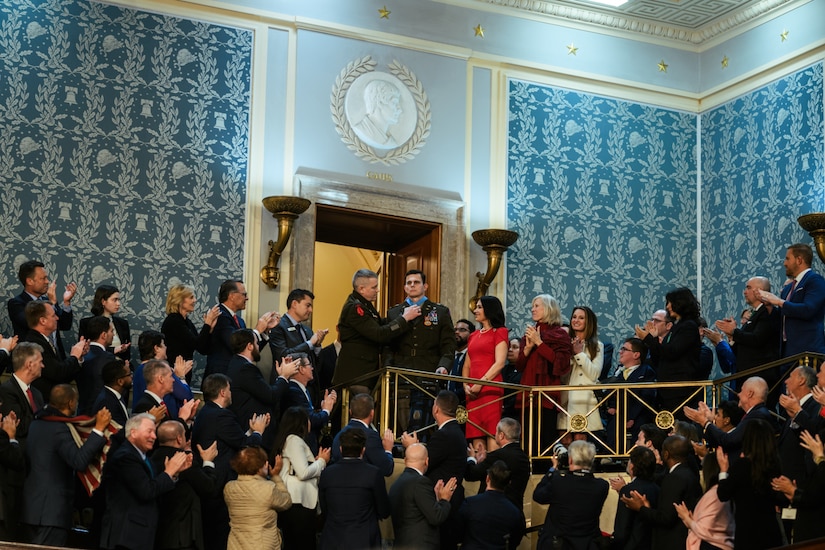 In the middle of a crowd room, a man in a military dress uniform places a medal around the neck of another man in similar attire. Dozens of people in business attire are clapping in the room.