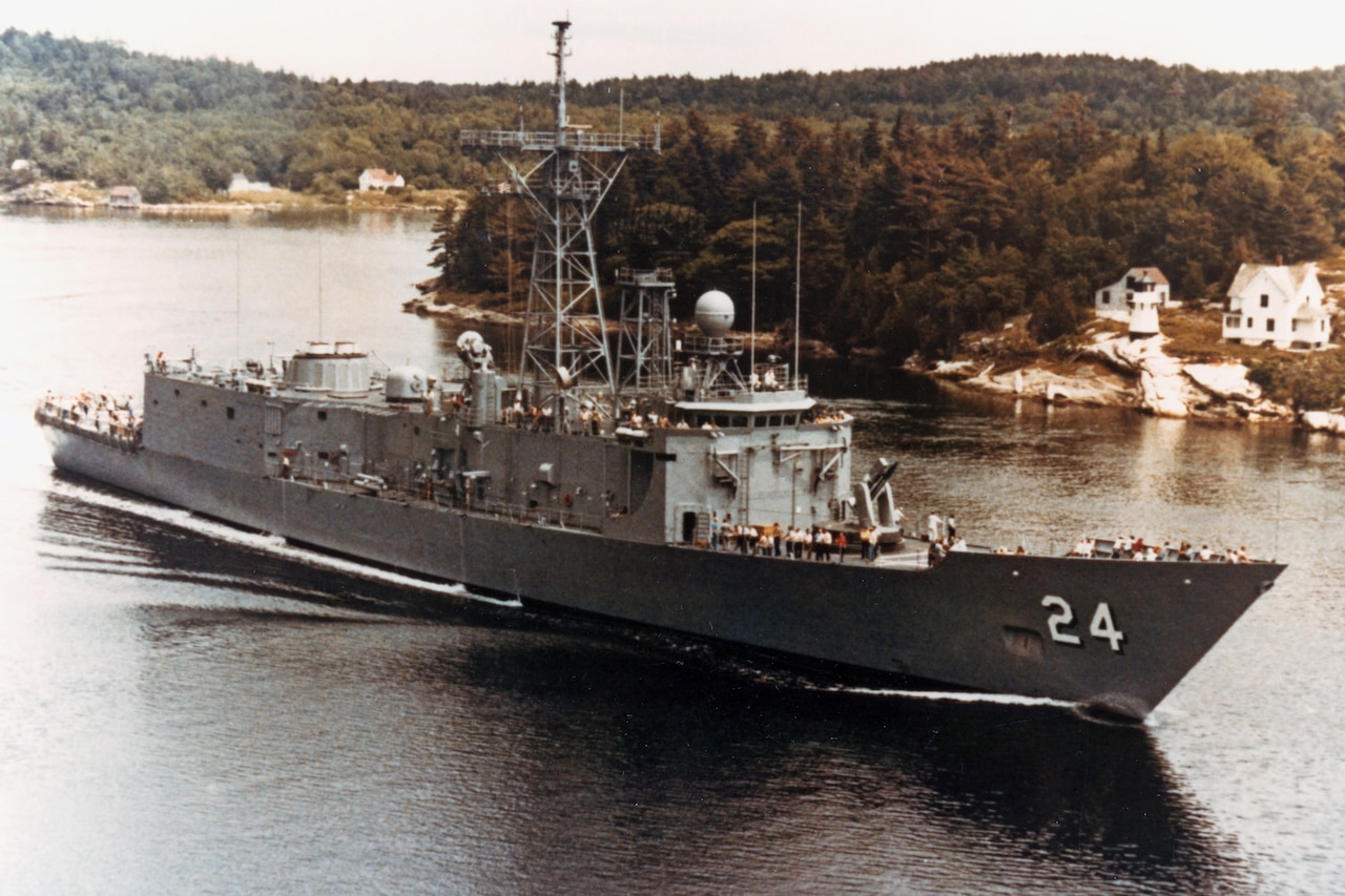 A large ship with dozens of people on the deck moves through water past forested land.