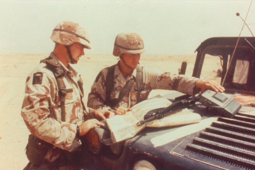 Two men wearing military camouflage uniforms stand in the desert and look at an electronic device and a map that is open on the hood of a vehicle.
