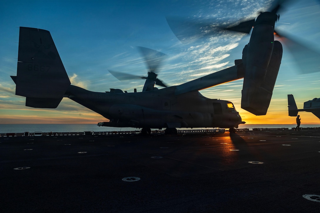 A military aircraft with propellers whirling takes off from the flight deck of a ship at sea during sunset.
