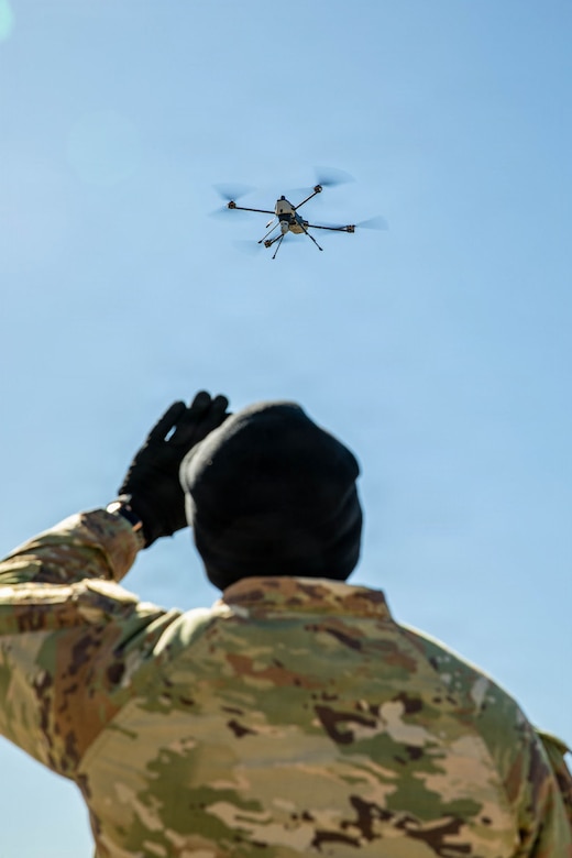 A man wearing a camouflage military uniform looks up at a drone flying in the air.