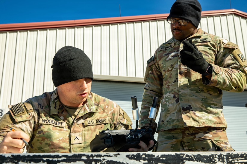 A man wearing a camouflage military uniform stands outside while looking down at another man in similar attire as he is seated holding a remote control. There is a building in the background.