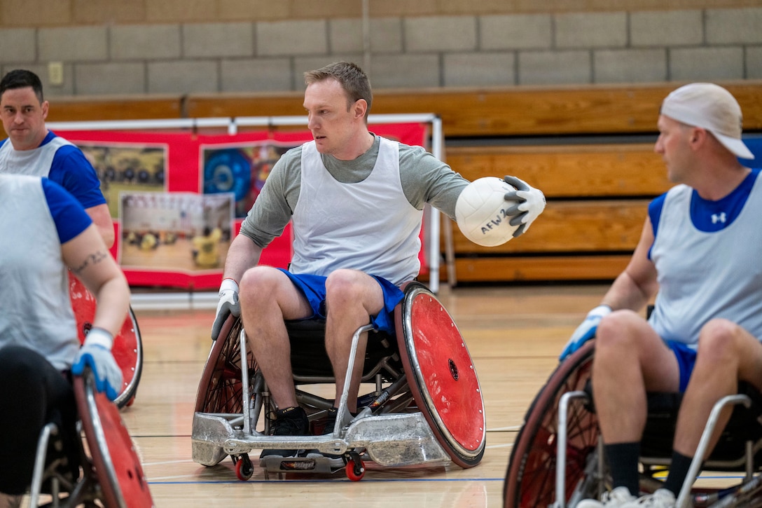 Four people using wheelchairs and wearing sports attire and gloves play rugby in a gymnasium.