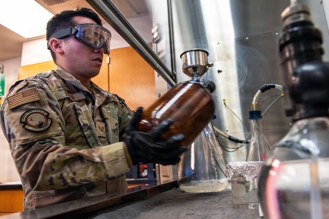 A man wearing a camouflage military uniform, gloves and safety goggles holds a brown glass bottle in a lab; other equipment is in the foreground.
