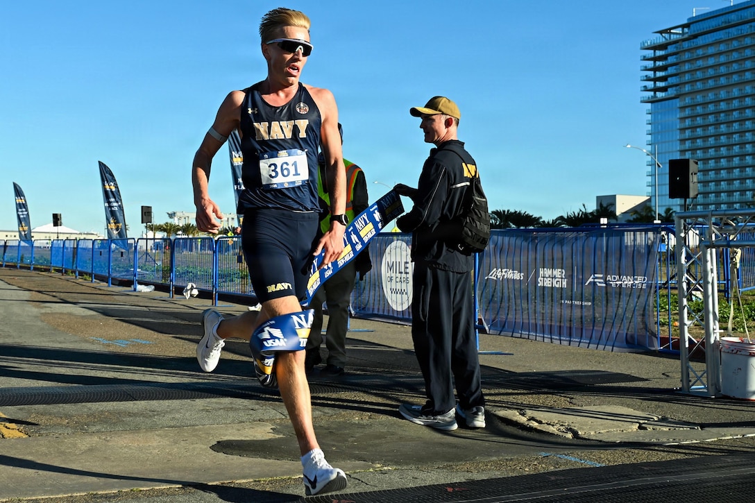 A man wearing sports attire and sunglasses runs through a racing tape held by a man to the right and an off-screen person, outside under a blue sky; an office building is in the background on the right.