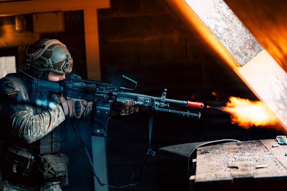 A person wearing a camouflage military uniform, helmet and gloves holds a weapon and looks through the scope in a dark area, while yellow, red and white flames emit from the nozzle.
