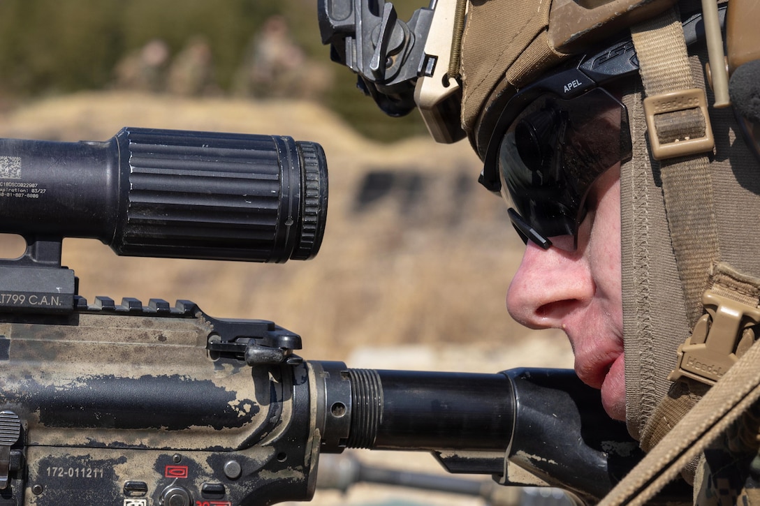 A man wearing a camouflage military uniform, helmet and sunglasses holds a weapon and looks through the scope outside in desert terrain.