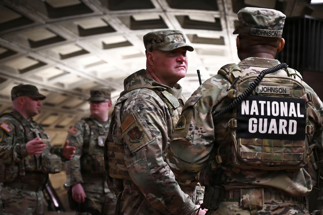 Four people in camouflage uniforms and hats stand and talk inside a train station tunnel; the person in the foreground wears a military vest with “National Guard” written on the back.