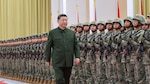 President Xi Jinping, wearing a dark green Mao-style suit, walks past rows of uniformed soldiers standing at attention during a formal indoor military inspection. The troops, dressed in camouflage uniforms and helmets, hold rifles across their chests in tight formation. Flags line the background of the large ceremonial hall.