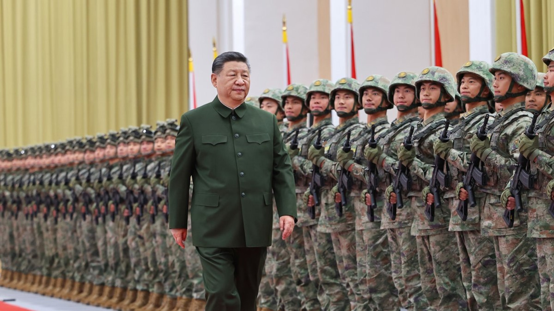 President Xi Jinping, wearing a dark green Mao-style suit, walks past rows of uniformed soldiers standing at attention during a formal indoor military inspection. The troops, dressed in camouflage uniforms and helmets, hold rifles across their chests in tight formation. Flags line the background of the large ceremonial hall.