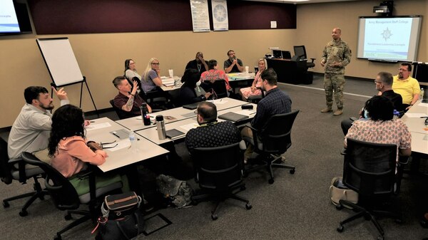 Students listen to an instructor in a military uniform in a classroom setting.