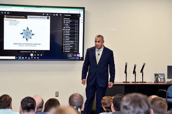 Professor lectures at the front of a classroom while a large group of students sit and listen.