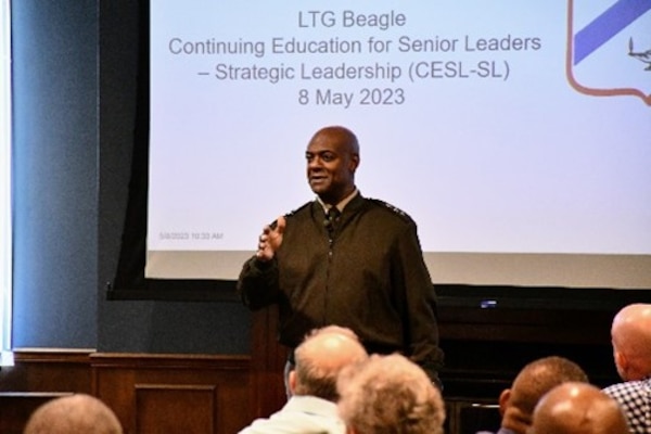 A man speaks and gestures while presenting to an audience in a conference room, with a slide behind him reading “LTG Beagle Continuing Education for Senior Leaders – Strategic Leadership (CESL-SL) 8 May 2023.