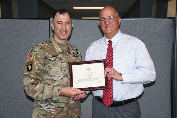 Two men, one in a U.S. Army uniform and one in a dress shirt and red tie, smile while holding a framed certificate together in an office setting.