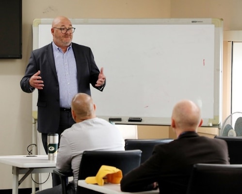 Civilian instructor stands in front of a whiteboard speaking to two adult students seated at tables in an Army Management Staff College classroom.