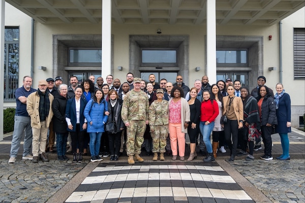 Group of civilian employees and Soldiers stand together outside a building entrance, with two Soldiers in uniform centered in the front row.