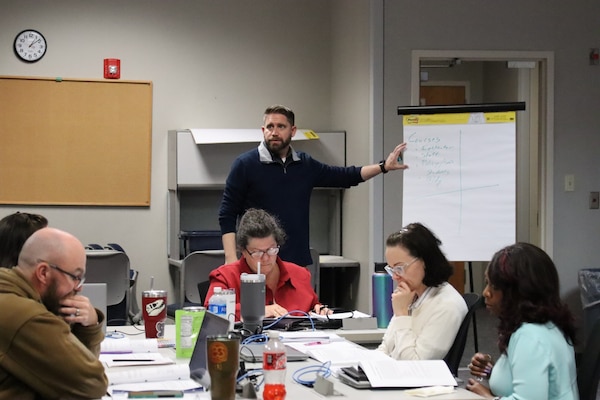 Instructor points to a flip chart while adult students seated at tables with laptops and papers participate in an Army Management Staff College classroom session.