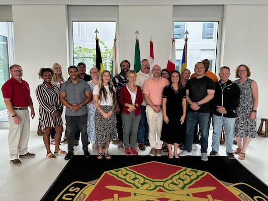 Group of adult students and staff stand on an Army Management Staff College emblem in a lobby with multiple national flags behind them.