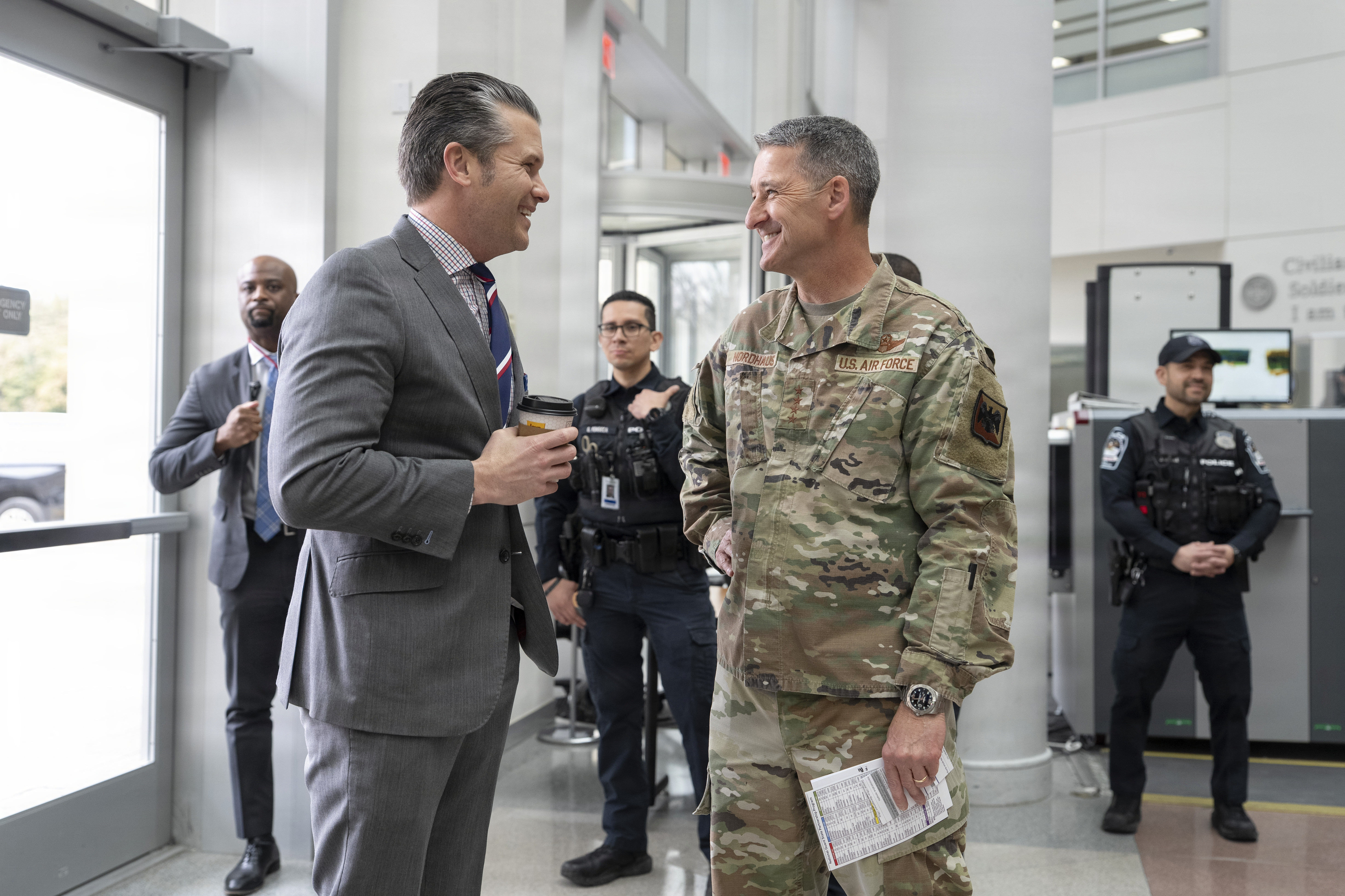 A man wearing a business suit smiles as he speaks with a man in a camouflage uniform. There are people dressed in police gear and civilian attire in the background. 