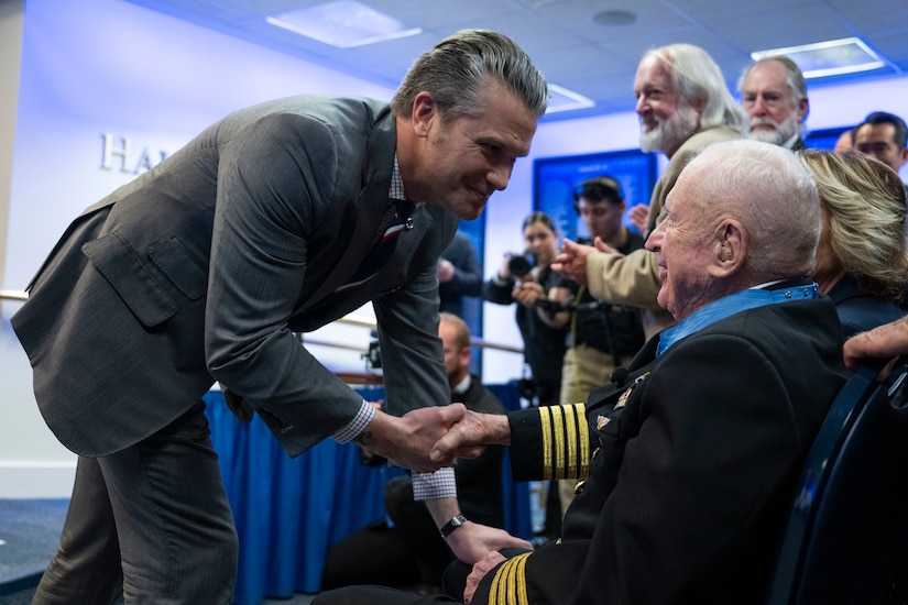 A man in business attire leans down to shake hands with a seated man in a formal military uniform; a crowd of people stands and claps inside a large ballroom.