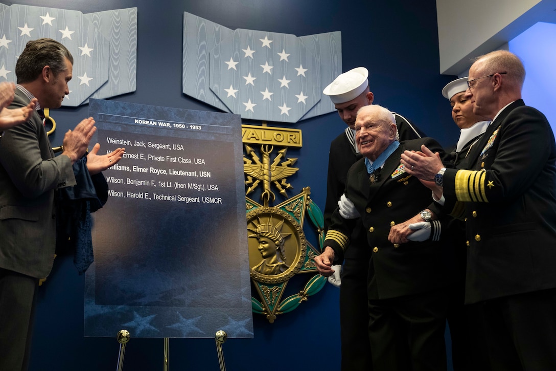 An older man in a formal military uniform smiles and stands with the assistance of another man in similar attire, as people in business attire and formal military uniforms clap. On the left of the man is a large sign displayed on an easel with the names of several service members.