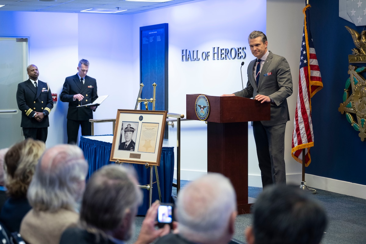 A man in business attire stands at a lectern and talks to an audience in a ballroom. In front of him is an easel with a framed picture displayed on it, and behind him is the American flag.