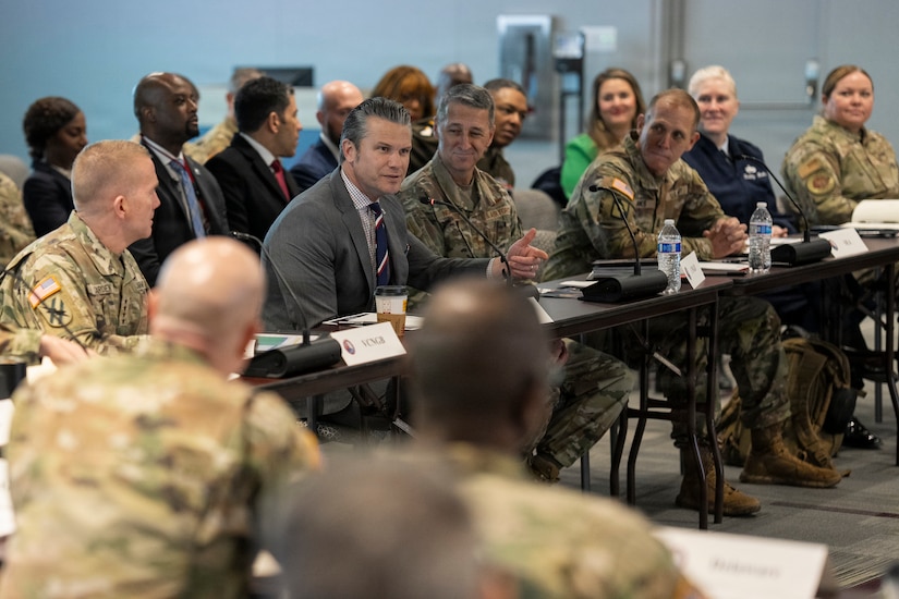 A man wearing a business suit sits at a table surrounded by people, also seated, dressed in camouflage military uniforms; one woman is wearing a blue military dress uniform. There are people in business attire seated in the background.