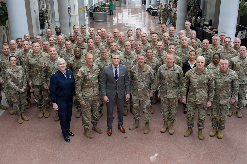 A man wearing a business suit poses for a photo with a group of people, mostly dressed in camouflage military uniforms. There is also a woman dressed in a blue military dress uniform and another woman in civilian attire.
