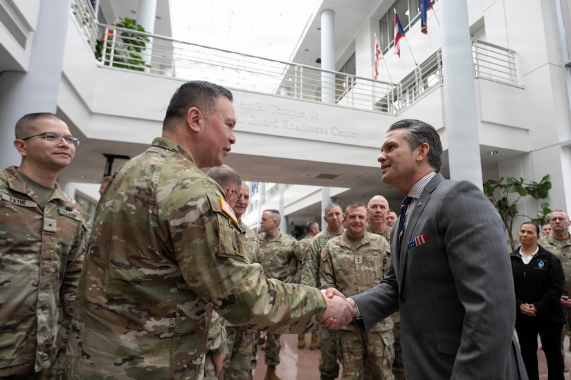 A man dressed in a business suit shakes hands with a man wearing a camouflage military uniform. There are people in camouflage military uniforms surrounding them.