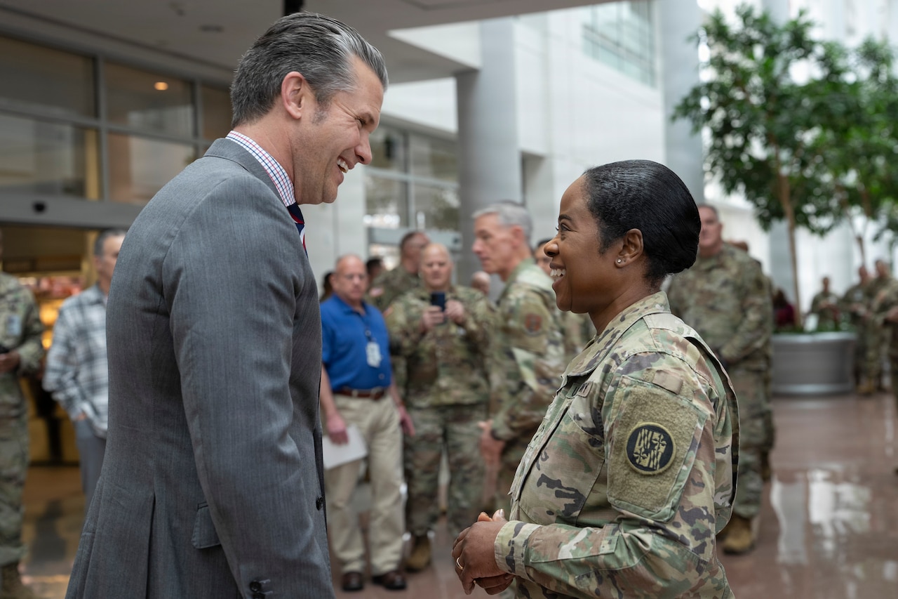 A man wearing a business suit smiles as he converses with a woman, who is also smiling, dressed in a camouflage military uniform.