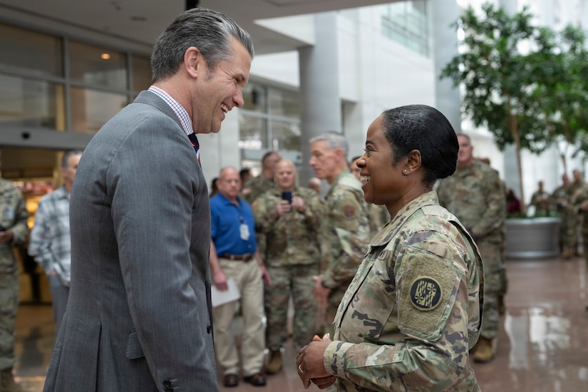 A man wearing a business suit smiles as he converses with a woman, who is also smiling, dressed in a camouflage military uniform.