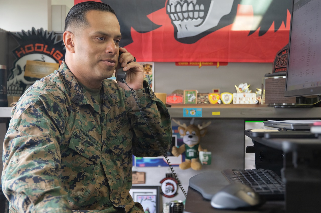 A Marine sitting behind a desk with computer equipment in an office setting holds a phone to his ear and speaks.