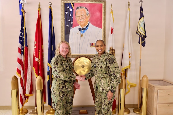 TWENTYNINE PALMS, Calif. (Feb. 19, 2026) — Lt. Gillian Gutierres, left, receives a Navy Medicine Readiness and Training Command Twentynine Palms challenge coin from commanding officer Capt. Janiese Cleckley after completing the Commanding Officer for a Day program. The presentation took place in front of a portrait of the hospital’s namesake, Medal of Honor recipient Robert E. Bush. (U.S. Navy photo by Christopher Jones, NMRTC Twentynine Palms public affairs officer)