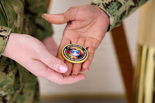 TWENTYNINE PALMS, Calif. (Feb. 19, 2026) — Lt. Gillian Gutierres  receives a Navy Medicine Readiness and Training Command Twentynine Palms challenge coin from commanding officer Capt. Janiese Cleckley after completing the Commanding Officer for a Day program. The presentation took place in front of a portrait of the hospital’s namesake, Medal of Honor recipient Robert E. Bush. (U.S. Navy photo by Christopher Jones, NMRTC Twentynine Palms public affairs officer)