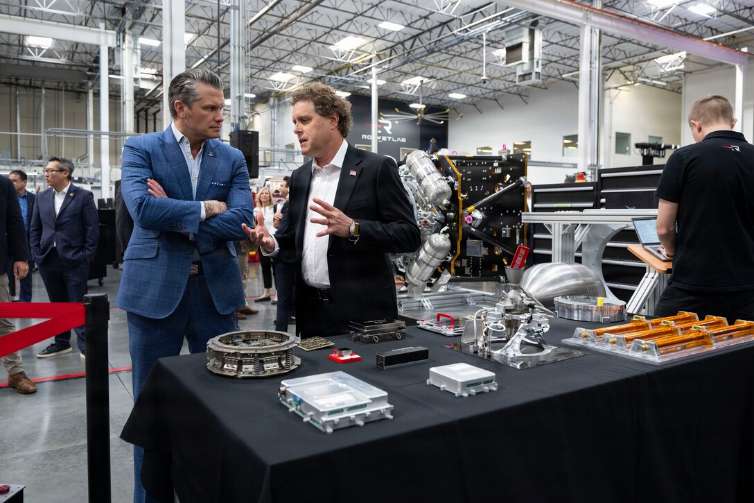 A civilian stands and speaks to Secretary of War Pete Hegseth in front of a display of equipment on a table.