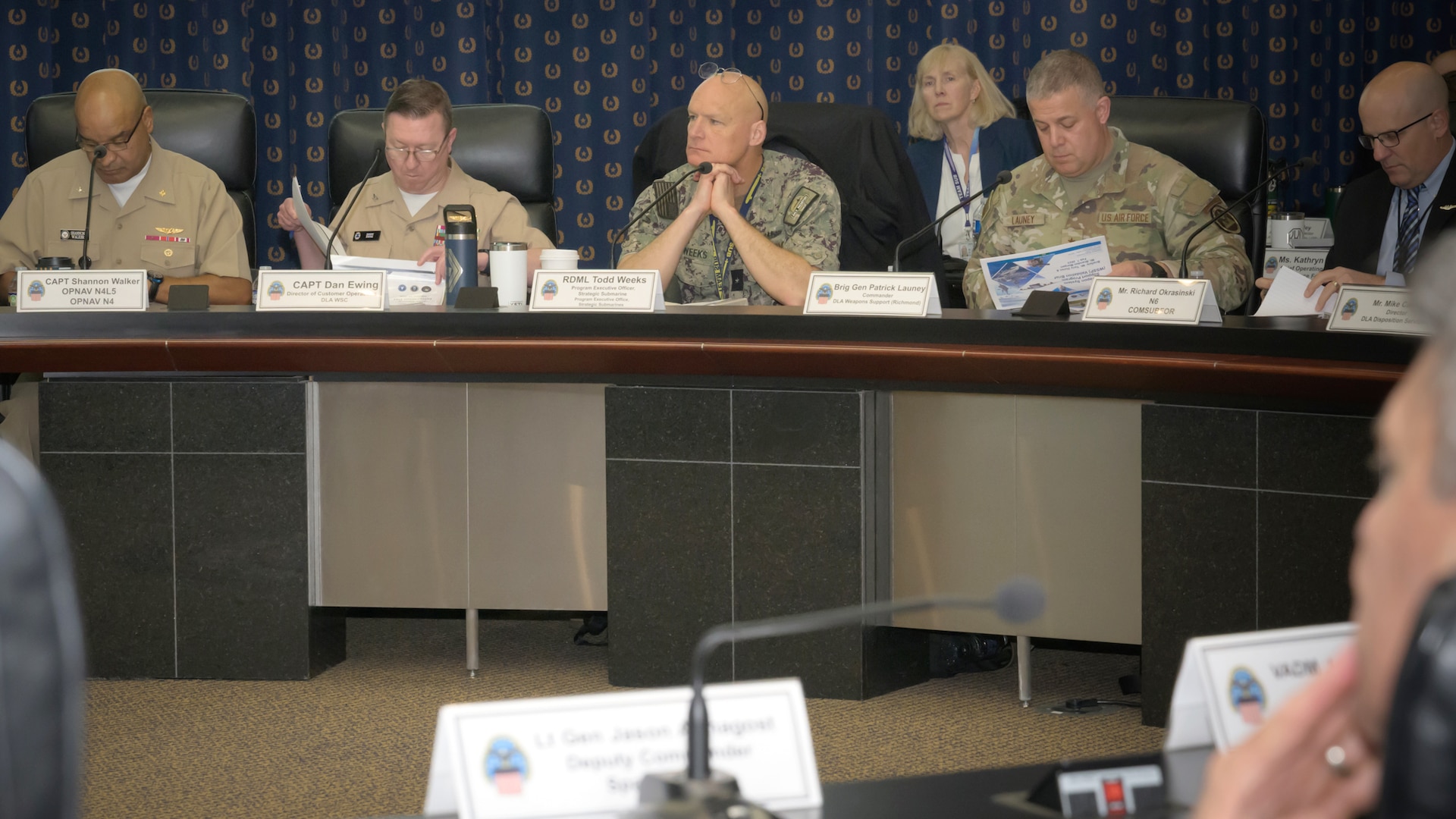 A panel of military officers and civilians are seated at a long conference table during a formal event. The participants are looking at documents, except for one man in the center who is listening with his hands clasped.