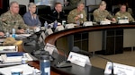 A group of military officers from various branches and a civilian woman are seated at a long, curved conference table during a formal meeting. They are listening intently, with one officer in the center speaking while holding a coffee mug.