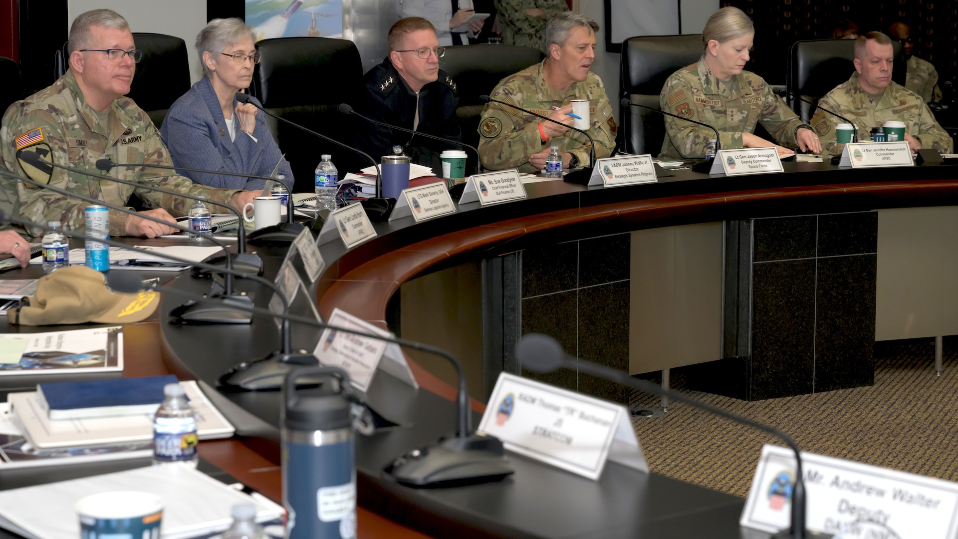 A group of military officers from various branches and a civilian woman are seated at a long, curved conference table during a formal meeting. They are listening intently, with one officer in the center speaking while holding a coffee mug.