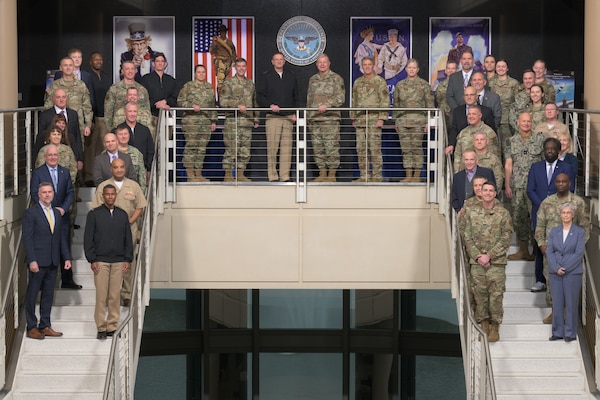 A large group of military personnel and civilians pose for a formal group picture on a multi-level staircase. They are arranged on a central landing and on the stairs to the left and right with a Department of Defense seal displayed between large, framed military-themed posters.