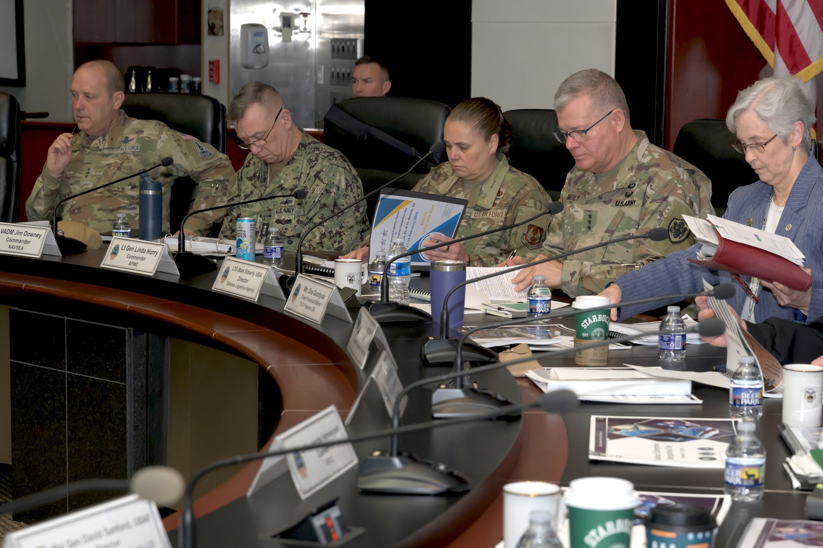 A group of military personnel in various uniforms and a woman in a blue suit are seated at a long, curved conference table during a formal meeting. They are all looking down at papers and binders in front of them.