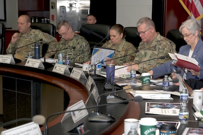A group of military personnel in various uniforms and a woman in a blue suit are seated at a long, curved conference table during a formal meeting. They are all looking down at papers and binders in front of them.