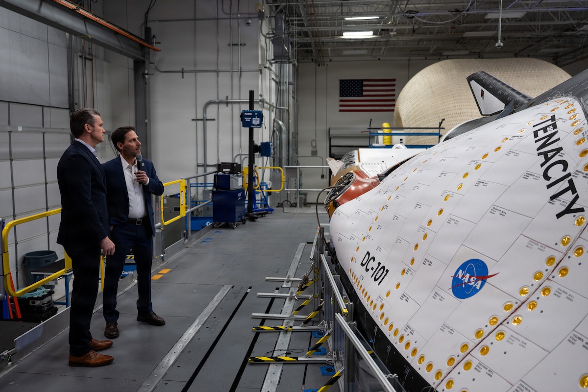 Secretary of War Pete Hegseth and another civilian view a large piece of equipment featuring a NASA logo in a factory-type setting.