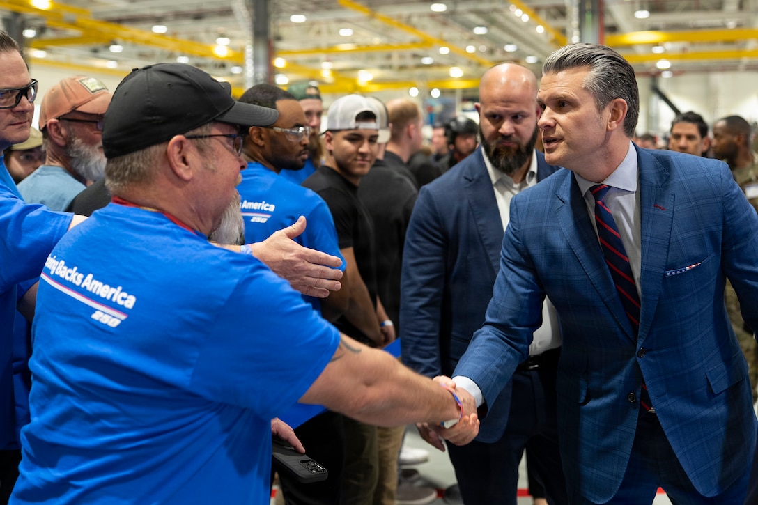 Secretary of War Pete Hegseth shakes hands with a civilian in a blue T-shirt amid a crowd in a large room.
