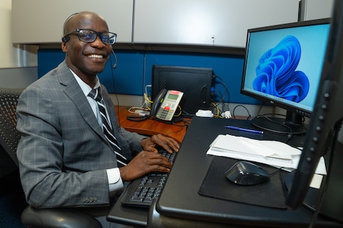 A man sits at a desk wearing a headset to answer phone calls, with his hands resting on a keyboard for his computer station.