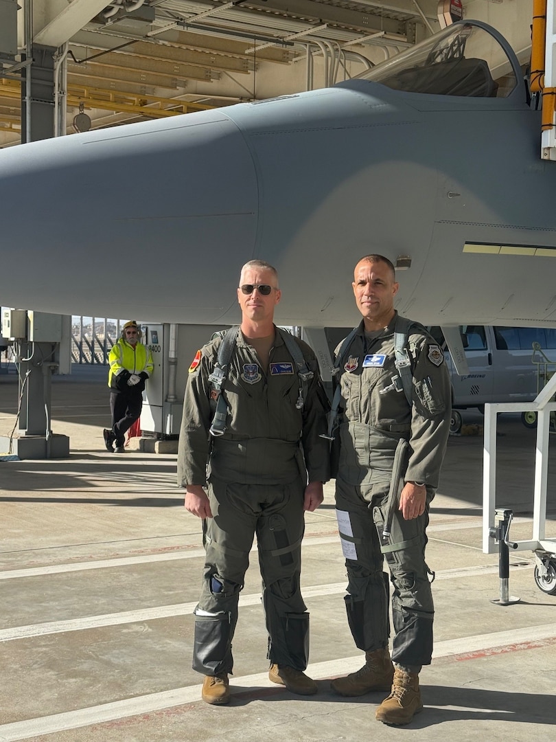 Two men wear flight suits and stand in front of an airplane